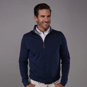 A man with short brown hair and a beard smiles while wearing the COLLARS AND CO - Heather Cotton Cloud 1/4 Zip Sweater in Navy with Grey Trim over a white collared shirt and cream pants, standing against a plain gray background.