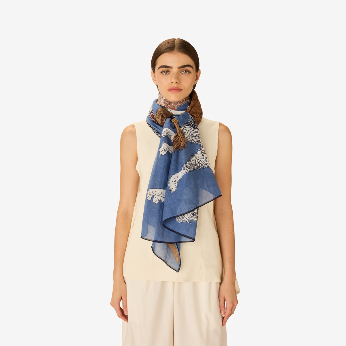 A person wears a sleeveless beige top and the INOUI EDITIONS - TOUTOU SCARF IN BLUE by INOUI, featuring a detailed animal print with white fur and brown accents, wrapped around their neck against a plain white background.