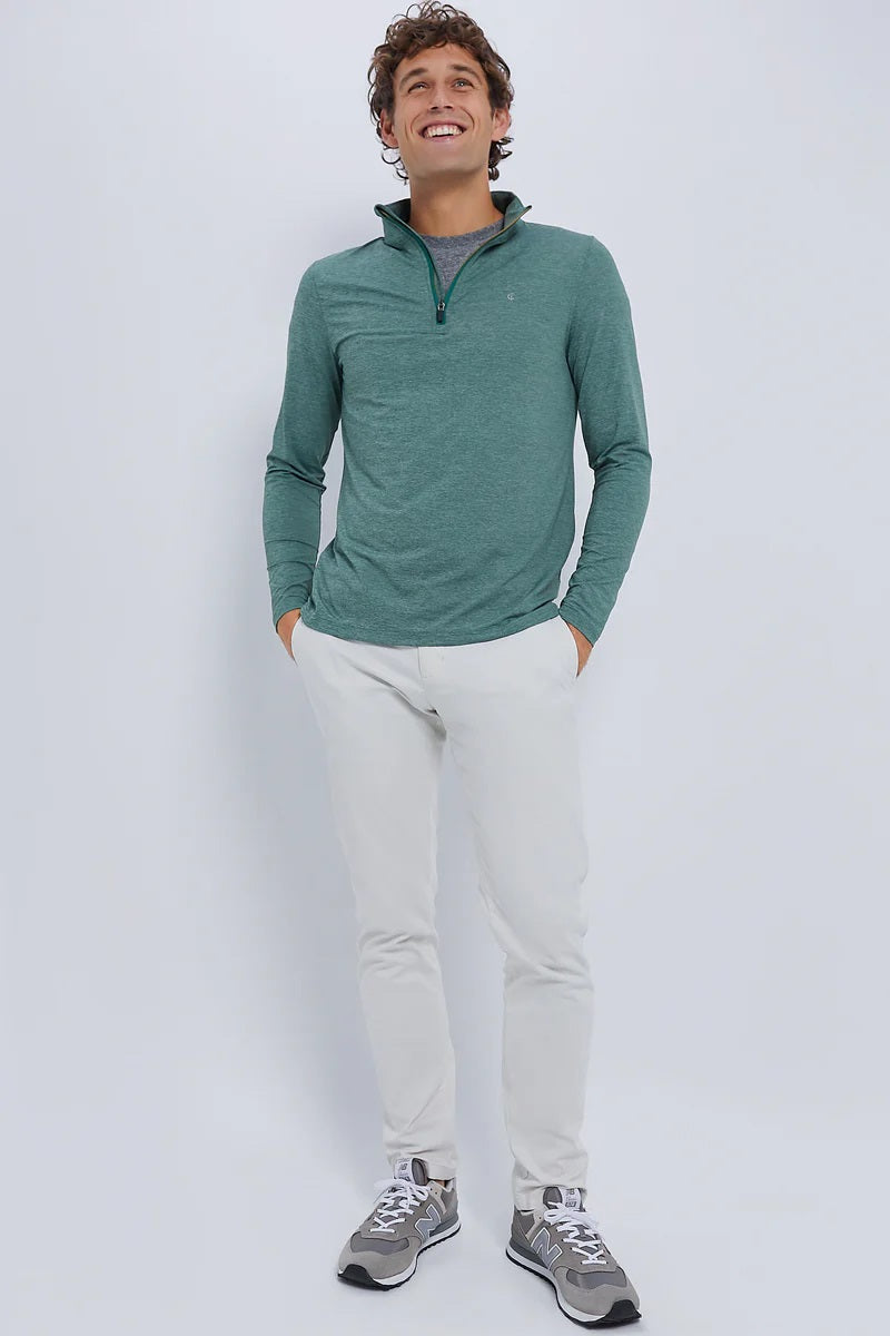 A man smiles against a plain light background, wearing the CRIQUET Feather Performance Knit Quarter Zip Pullover in Forest Heather by CRIQUET, paired with white pants and gray sneakers. His hands are in his pockets and he looks relaxed and cheerful.