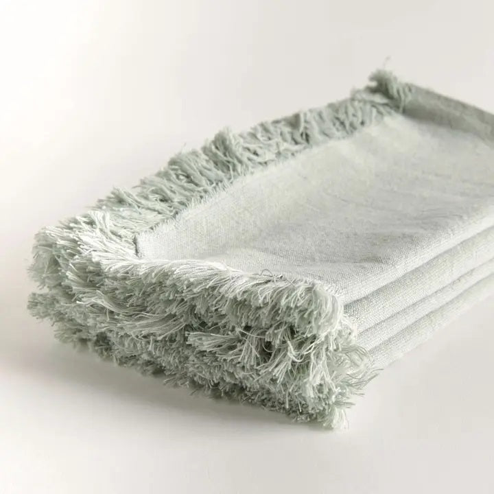 A stack of NAPA HOME AND GARDEN - AURA DINNER NAPKIN IN LIGHT GREEN, featuring fringed edges, is folded and placed on a white surface.