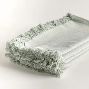 A stack of NAPA HOME AND GARDEN - AURA DINNER NAPKIN IN LIGHT GREEN, featuring fringed edges, is folded and placed on a white surface.
