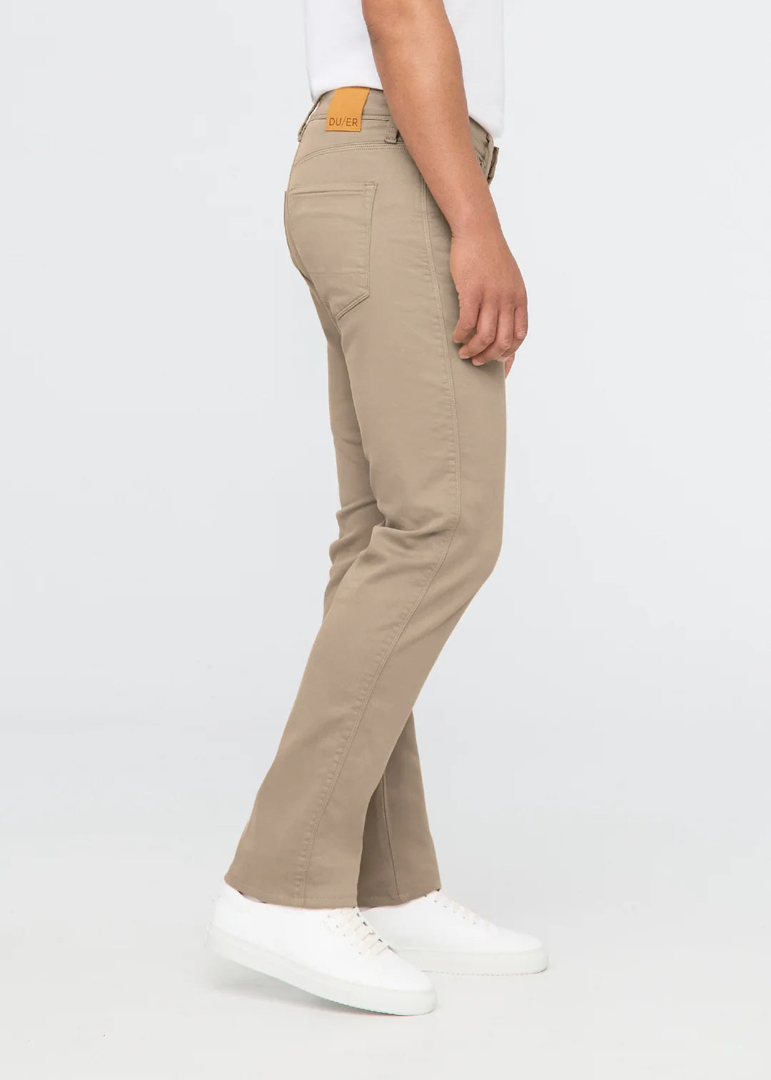 A person wearing DUER NO SWEAT STRAIGHT PANTS IN DESERT KHAKI, a white t-shirt, and white sneakers stands against a plain light gray background.