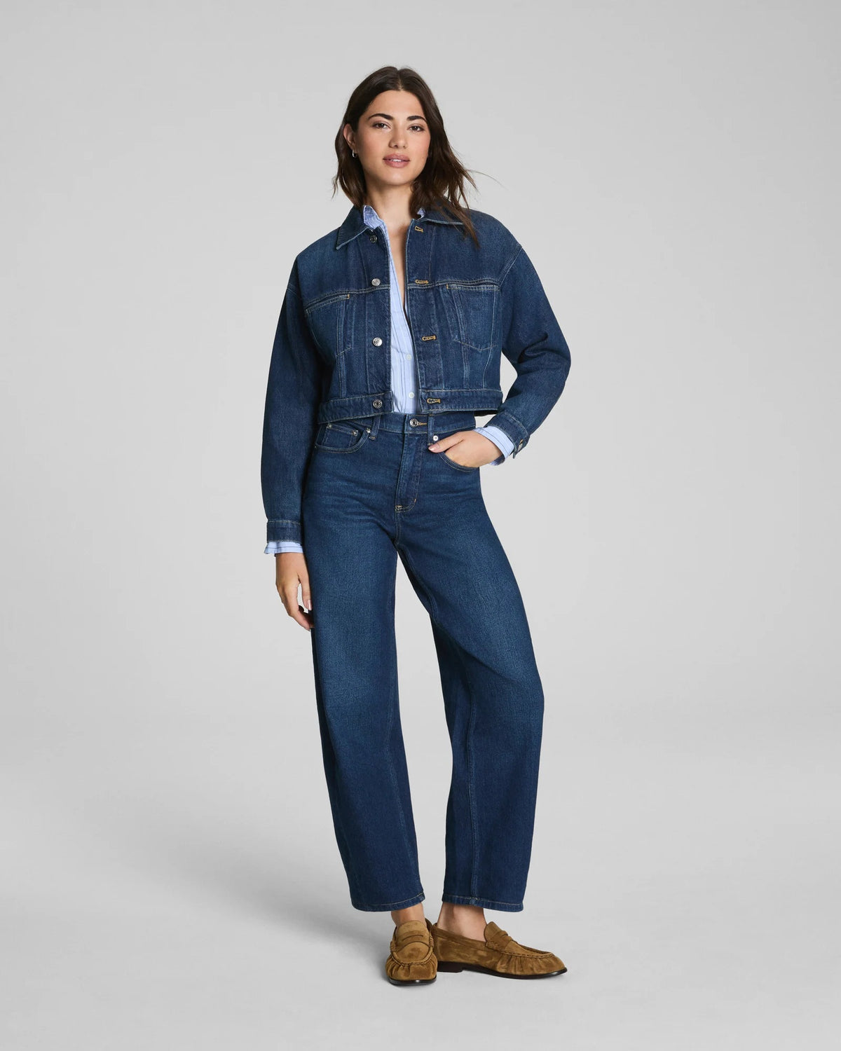 Here is the rewritten sentence using your product data: A woman with long dark hair and a relaxed expression stands against a plain background wearing the SPANX SPANXSHAPE Authentic 360 Barrel Leg Jean in Adriatic Blue, paired with a matching blue denim jacket, light blue shirt, and brown suede loafers.