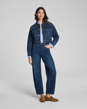 Here is the rewritten sentence using your product data: A woman with long dark hair and a relaxed expression stands against a plain background wearing the SPANX SPANXSHAPE Authentic 360 Barrel Leg Jean in Adriatic Blue, paired with a matching blue denim jacket, light blue shirt, and brown suede loafers.