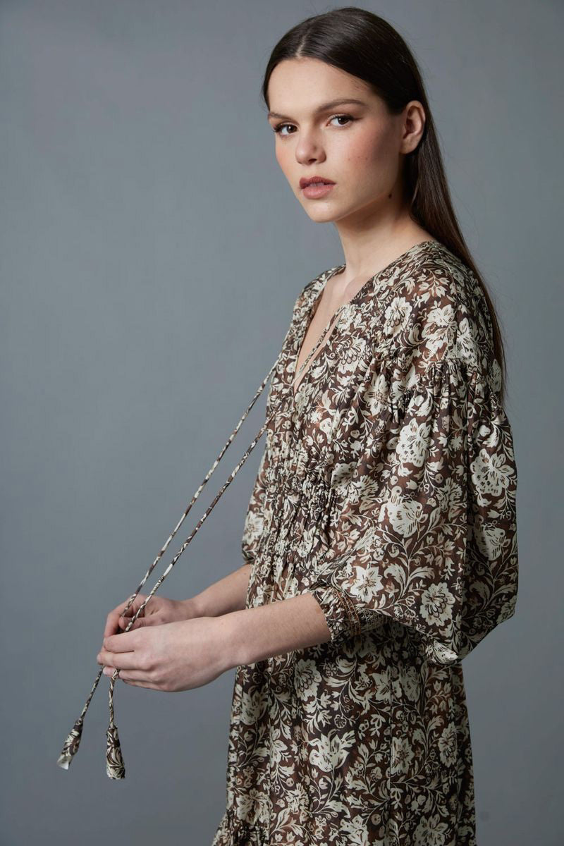 A woman stands against a gray background wearing the HOPE FOR FLOWERS TIERED PEASANT DRESS with puffed sleeves and a shirred bodice. She has her hands in its pockets and pairs dark loafers with light-colored socks.