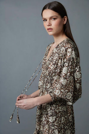 A woman stands against a gray background wearing the HOPE FOR FLOWERS TIERED PEASANT DRESS with puffed sleeves and a shirred bodice. She has her hands in its pockets and pairs dark loafers with light-colored socks.