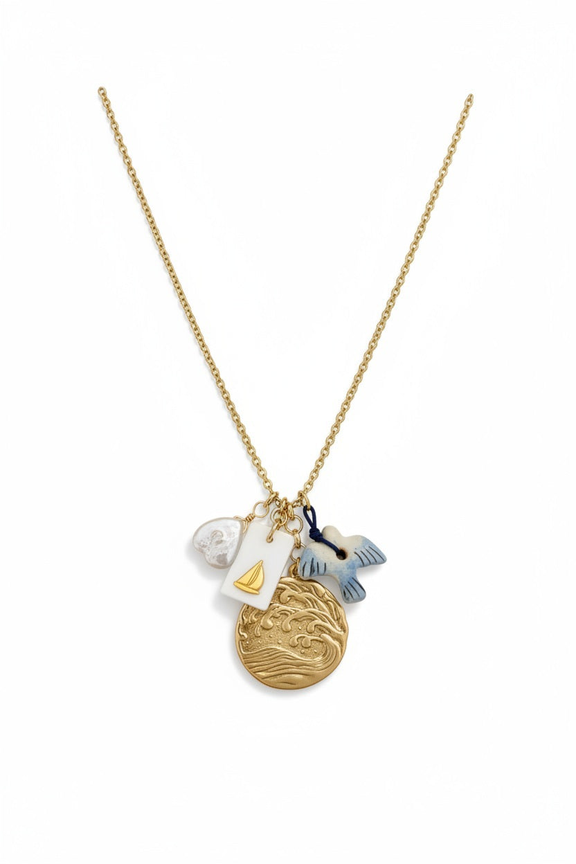 The SANDRINE FRANCE STUDIO WREN NECKLACE is a handmade gold necklace from NYC featuring four charms—a white shell, a rectangular gold sailboat tag, a gold wave pendant, and a blue-white fish—on a white jewelry stand.