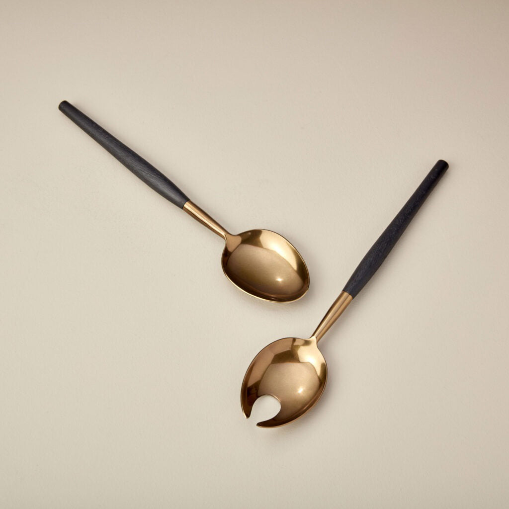 A round, deep bowl from BE HOME, INC&#39;s AGED BRONZE SERVING SET with a smooth, golden-bronze finish sits on a light wooden surface. The slightly irregular, organic shape of the bowl enhances its handcrafted appearance. The background displays the light wood grain pattern of the tabletop.