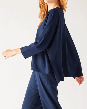 A woman with wavy brown hair models the MERSEA - CATALINA SLUB TEE—an oversized, long-sleeved white top with thin gray stripes—by MERSEA. She pairs it with blue jeans and stands against a plain, light background, hands behind her back.