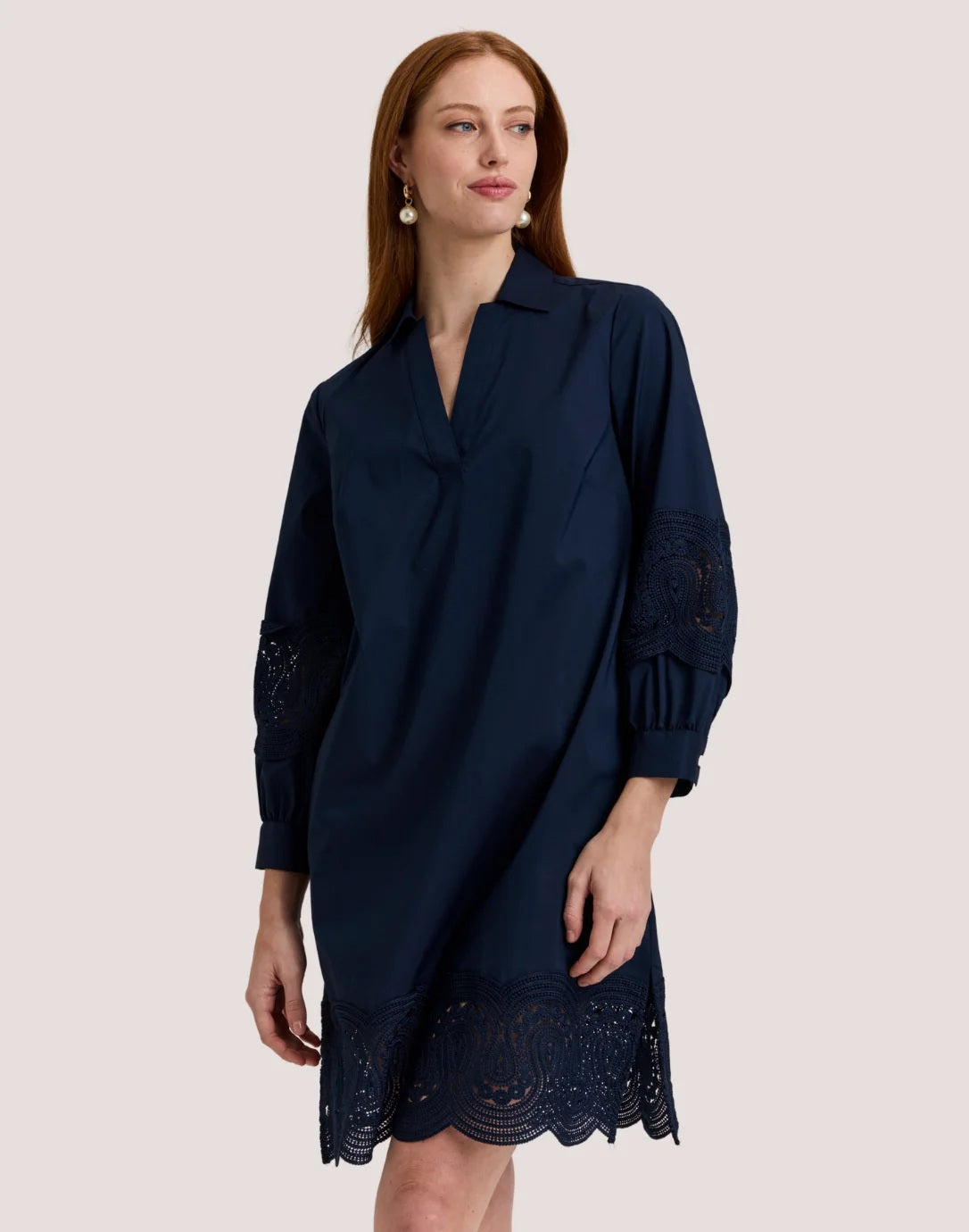 A woman with long red hair wears the HINSON WU Mona Bracelet Sleeve Lace Stretch Polished Cotton Dress in Navy. Crafted from stretch polished cotton, the dress features eyelet lace details on the sleeves and hem. She stands against a plain light background, looking slightly to the side with a neutral expression, her outfit highlighting its polished finish.
