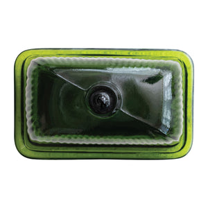The CREATIVE COOP GREEN PRESSED GLASS BUTTER DISH is displayed with green glass containers and bottles on a textured white surface, beside a folded towel and a partially visible book.