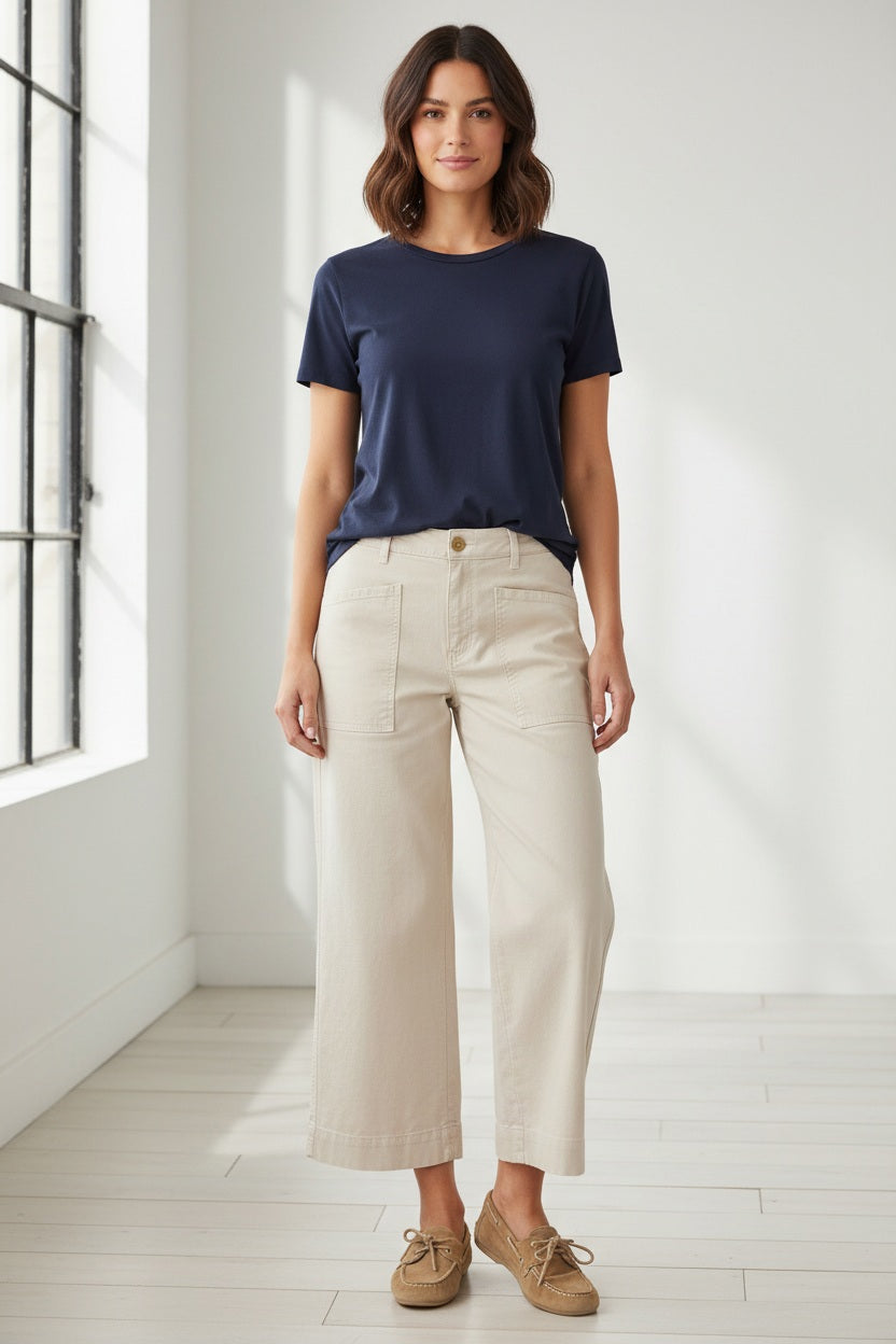 A person wearing the FRANK &amp; EILEEN Limerick Cropped Wide Leg Carpenter Pant in Italian Performance Linen Cement, styled with tan lace-up shoes, stands indoors on a wooden floor with wicker furniture in the background.