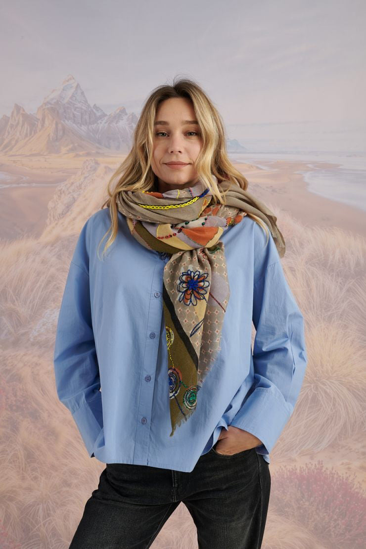 A woman with long blonde hair, in a light blue shirt and dark pants, smiles in front of a scenic backdrop, draping the elegant, hand-embroidered STORIATIPIC - ASTER WOOL SCARF around her shoulders.