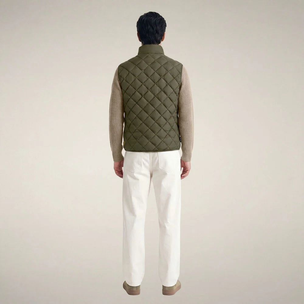A person stands facing forward, wearing the SAVE THE DUCK - FABRICE Puffer Vest in Snake Green with Plumtech® padding over a beige sweater, paired with white pants and tan shoes, against a plain light background.