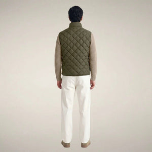 A person stands facing forward, wearing the SAVE THE DUCK - FABRICE Puffer Vest in Snake Green with Plumtech® padding over a beige sweater, paired with white pants and tan shoes, against a plain light background.