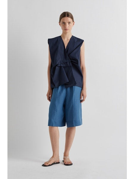 A person wears MEIMEIJ's Pull On Linen Short in Cerulean featuring a loose fit, knee length, and elastic waist, styled with flat strappy sandals against a plain light-colored background.