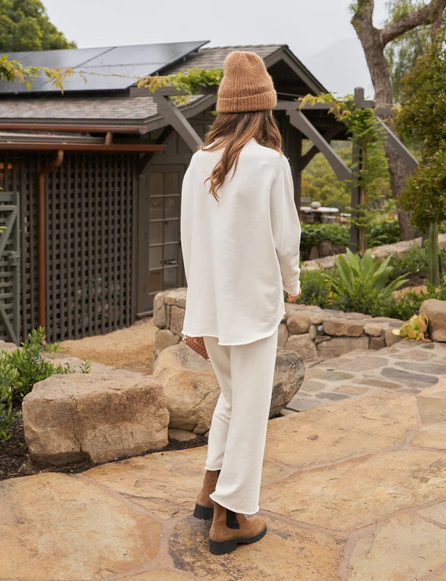 A woman with long brown hair stands smiling near a black grand piano in a modern, well-lit room. She wears the FRANK AND EILEEN - EFFIE LONG SLEEVE FUNNEL NECK CAPELET from FRANK &amp; EILEEN paired with blue jeans. A large window and a lamp are in the background, enhancing the cozy winter warmth of her stylish ensemble.