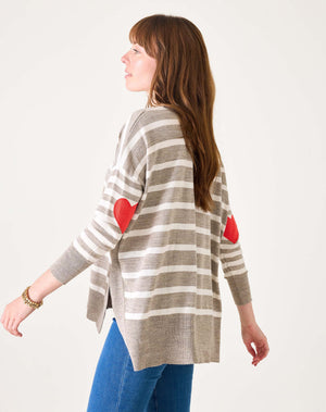 A woman with long brown hair in blue jeans models the MERSEA - AMOUR SWEATER IN DRIFTWOOD STRIPE by MERSEA, featuring gray and white stripes with red heart-shaped elbow patches. She stands sideways against a plain white background.
