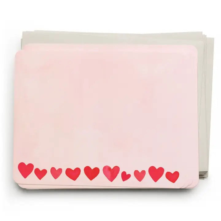 The E. FRANCES - FLAT NOTES BLUSH MINI HEARTS by E. FRANCES PAPER are pale pink notecards with rounded corners and red and pink hearts along the bottom, stacked atop light gray envelopes.
