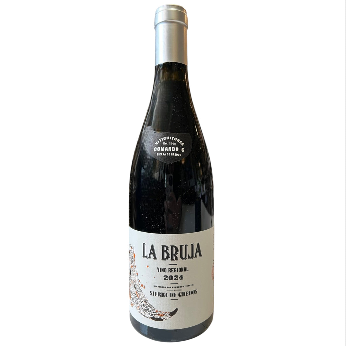 A bottle of COMANDO G 'La Bruja' Grenache 2024 by Wine: Freedom Beverage stands on a counter before shelves of other wines. The bottle features a white label with black and red text.
