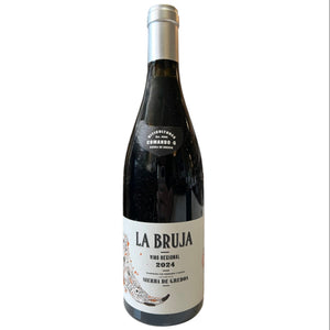 A bottle of COMANDO G 'La Bruja' Grenache 2024 by Wine: Freedom Beverage stands on a counter before shelves of other wines. The bottle features a white label with black and red text.