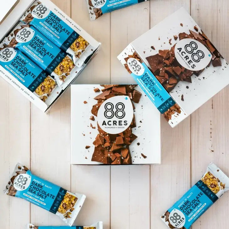 Flat lay of 88 ACRES - DARK CHOCOLATE SEA SALT SEED OAT BAR, a nut-free vegan snack, featuring opened and unopened bars in blue wrappers arranged with white boxes and chocolate pieces on a light wooden surface.