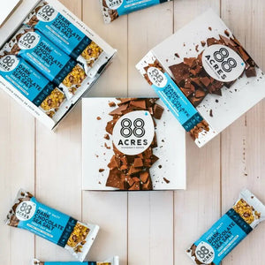 Flat lay of 88 ACRES - DARK CHOCOLATE SEA SALT SEED OAT BAR, a nut-free vegan snack, featuring opened and unopened bars in blue wrappers arranged with white boxes and chocolate pieces on a light wooden surface.