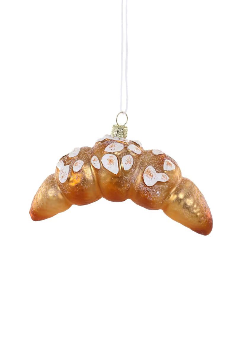 The ALMOND CROISSANT ORNAMENT from CODY FOSTER is a glossy glass Christmas decoration shaped like a croissant. It hangs by a white string, featuring intricate icing details that resemble powdered sugar for an added touch of whimsy.