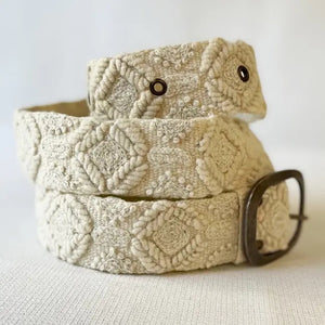 A person sits outdoors on stone pavement, shown from torso to lap, wearing a light green t-shirt, beige pants, and the JENNY KRAUSS - WANDERING CLOUDS EMBROIDERED BELT with a decorative buckle. Head and lower legs are not visible.