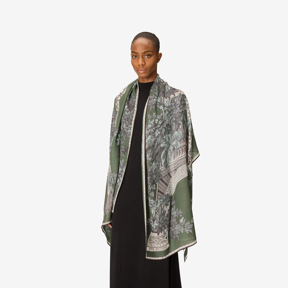 A person with short hair wears a long black dress and the INOUI EDITIONS 1889 WOOL SCAR FIN KHAKI scarf by INOUI—a green patterned accessory featuring intricate floral and geometric designs, crafted from 100% wool in India—while standing against a plain white background.