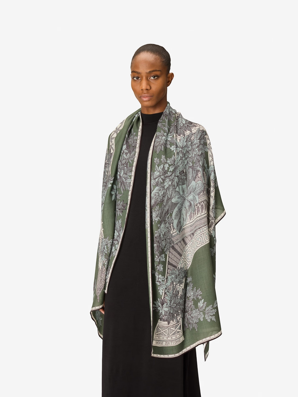 A person with short hair wears a long black dress and the INOUI EDITIONS 1889 WOOL SCAR FIN KHAKI scarf by INOUI—a green patterned accessory featuring intricate floral and geometric designs, crafted from 100% wool in India—while standing against a plain white background.