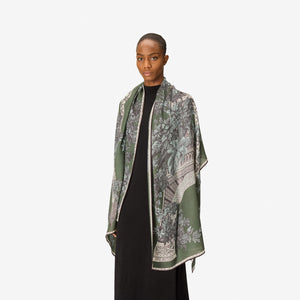 A person with short hair wears a long black dress and the INOUI EDITIONS 1889 WOOL SCAR FIN KHAKI scarf by INOUI—a green patterned accessory featuring intricate floral and geometric designs, crafted from 100% wool in India—while standing against a plain white background.