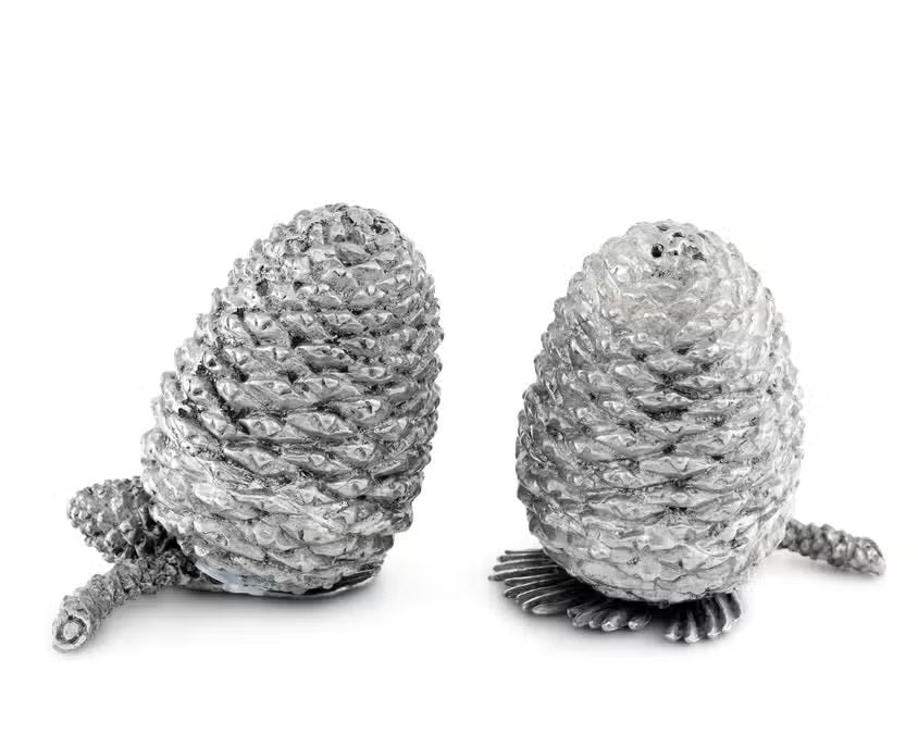 Two detailed silver pine cones stand upright on a white background, resembling the VAGABOND HOUSE - PEWTER PINE CONESALT AND PEPPER SET by VAGABOND HOUSE, with small pine needle segments visible at their bases.