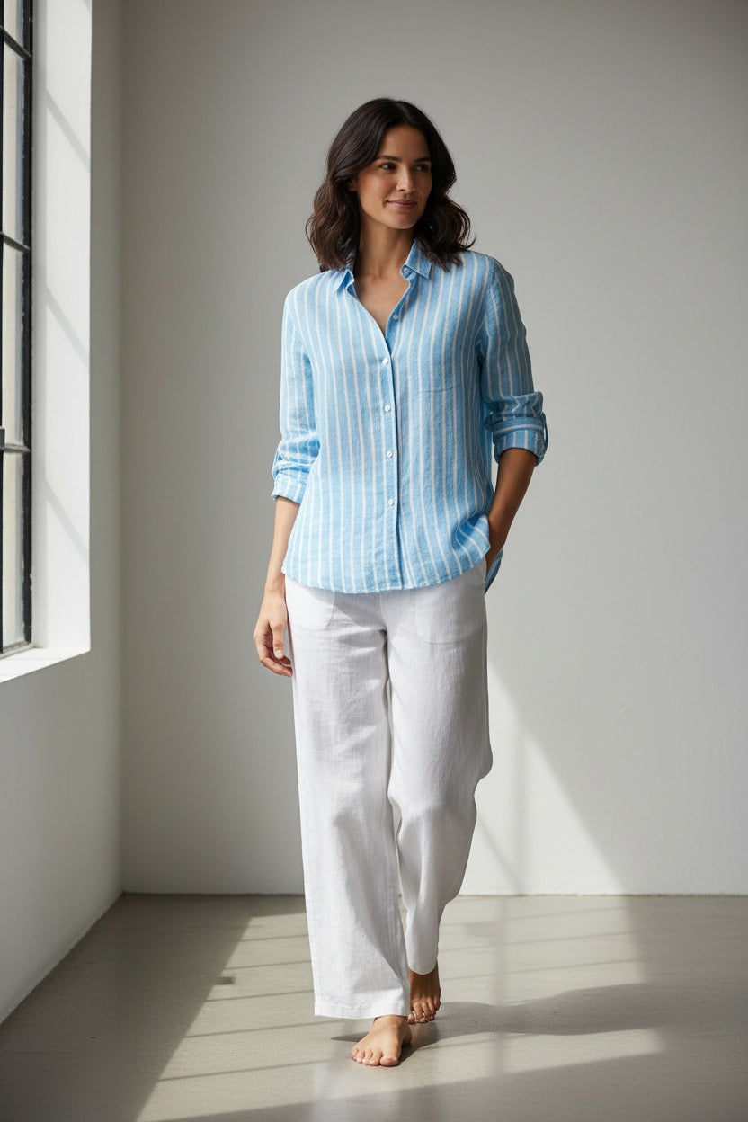 A woman stands outside near the beach, smiling. She wears the FRANK &amp; EILEEN EILEEN Relaxed Button Up Shirt in Italian Linen Turquoise Stripe and white pants, with her hands in her pockets.