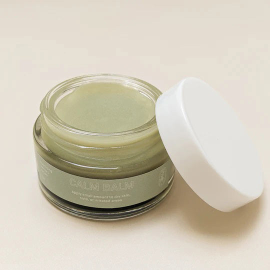 A small glass jar labeled "PINK HOUSE ORGANICS - CALM BALM" containing a light green, soothing balm. The jar is open, and the white lid is placed next to it on a neutral background. Ideal for all skin types, the balm appears smooth, indicating it hasn't been used yet.
