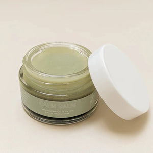 A small glass jar labeled "PINK HOUSE ORGANICS - CALM BALM" containing a light green, soothing balm. The jar is open, and the white lid is placed next to it on a neutral background. Ideal for all skin types, the balm appears smooth, indicating it hasn't been used yet.