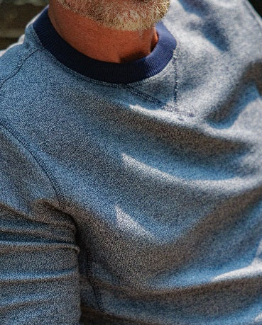 A man with short brown hair wearing the CRIQUET - French Terry Crewneck Sweatshirt in Dusty Blue and white pants stands with one hand in his pocket, looking to his left against a white background.