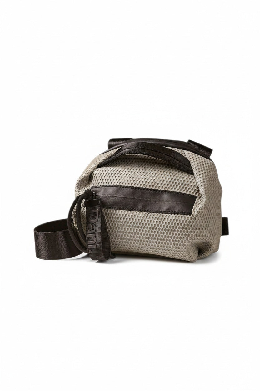 The DANIELLA LEHAVI BONNIE MINI TRAVEL CROSSBODY, a small brown vegan bag with black accents and strap, is displayed on a white pedestal against a plain beige background.