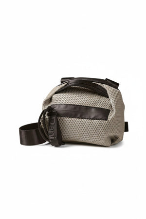 The DANIELLA LEHAVI BONNIE MINI TRAVEL CROSSBODY, a small brown vegan bag with black accents and strap, is displayed on a white pedestal against a plain beige background.