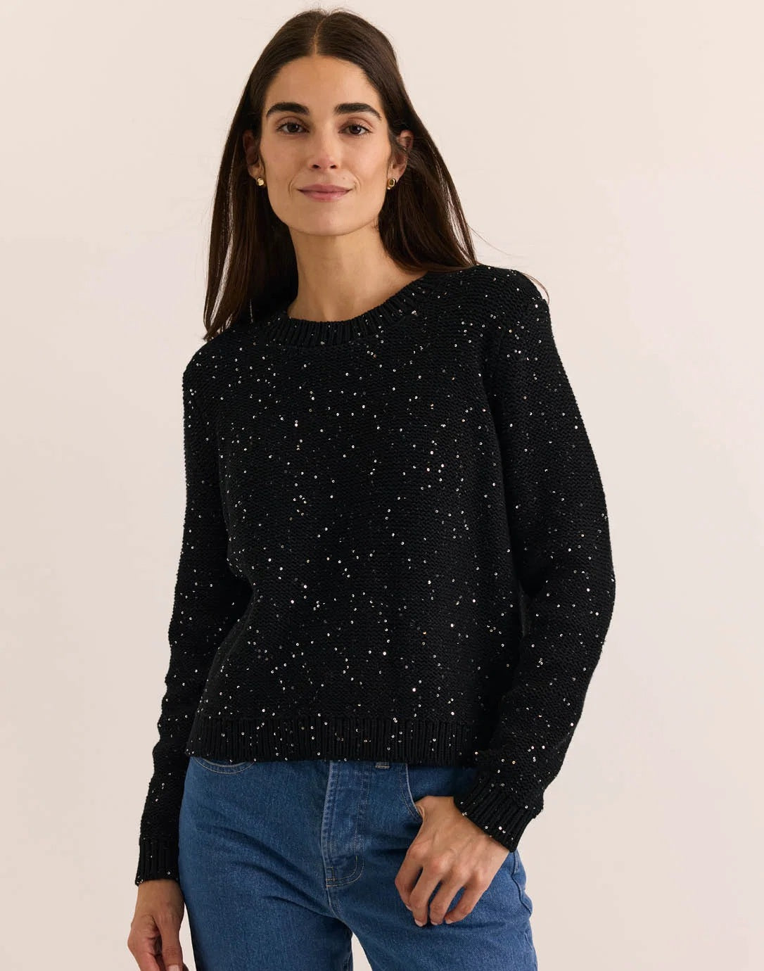 A woman with long dark hair, dressed in the HINSON WU - QUEENIE LONG SLEEVE SEQUIN SWEATER IN BLACK by HINSON WU and blue jeans, stands against a plain light background, smiling gently at the camera.