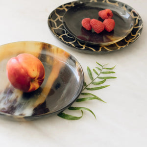 The SPLATTER HORN PLATE MEDIUM by BE HOME, INC is a round, shallow dish handcrafted in Vietnam from polished water buffalo horn, showcasing a marbled pattern of light brown, cream, and dark brown tones.