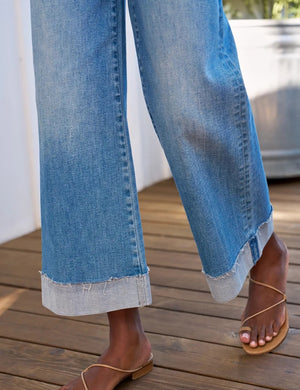 A person stands on a wooden deck with their back to the camera, wearing the FRANK & EILEEN WESTPORT Illusion Cuffed Hem Pull On Jean in 2013 Wash, styled with a white shirt and sandals. A large metal planter filled with greenery sits beside them.
