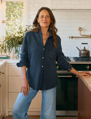 A woman with long brown hair, wearing the FRANK & EILEEN Eileen Relaxed Button Up Sweatshirt in Vintage Navy and light blue jeans, stands in a bright kitchen with white cabinets, a green stove, and a window filled with plants.