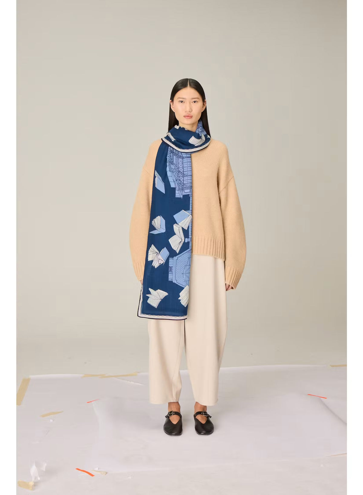 A woman stands against a plain background, wearing a beige sweater, cream wide-leg pants, black shoes, and the INOUI EDITIONS - VIVIENNE WOOL SCARF IN BLUE by INOUI—a long blue scarf with a white pattern made from 100% wool in India and measuring 100 x 190 cm.