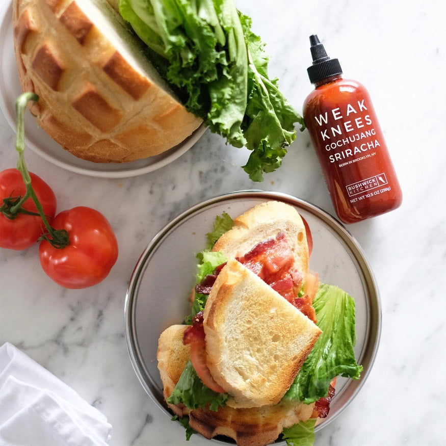 A 10.5 oz (298g) bottle of BUSHWICK KITCHEN - 'WEAK KNEES' GOCHUJANG SRIRACHA hot sauce features a striking red design with a black cap and white text, proudly made in Brooklyn, NY, USA. This unique blend includes fermented chile paste for an exceptional kick.