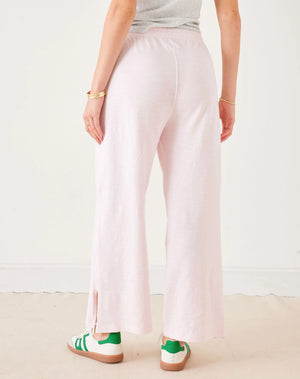 A woman stands in a minimal white room wearing the MERSEA TRAVELER TEE PANT, light gray, made from Turkish cotton slub—styled with white sneakers featuring green accents—and smiles softly at the camera.