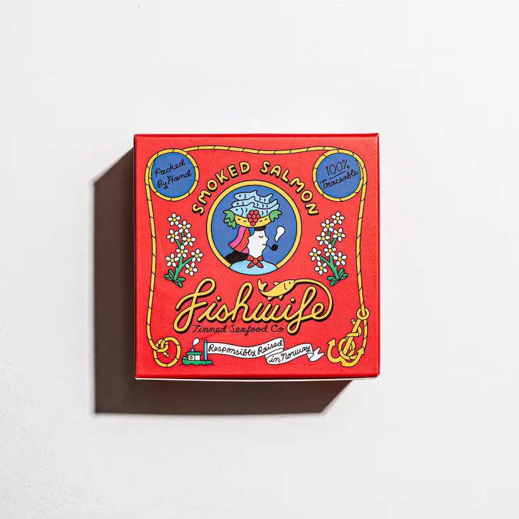 A vibrant red tin of FISHWIFE - SMOKED ATLANTIC SALMON features a colorful woman with fish and lemons, proudly stating "100% Traceable," "Responsibly Raised in Norway," and honoring sustainable seafood, from the brand FISHWIFE.