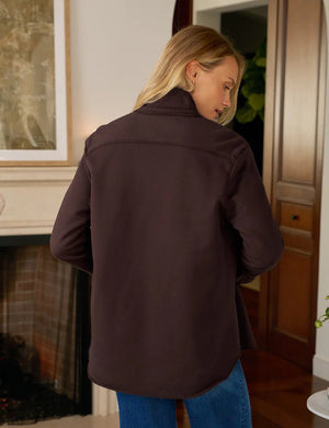 A woman with long blonde hair wears the FRANK AND EILEEN - EILEEN Relaxed Button Up Shirt in Triple Fleece Coco, by FRANK & EILEEN. Indoors, she looks down and adjusts her sleeve in a cozy, warmly lit room.