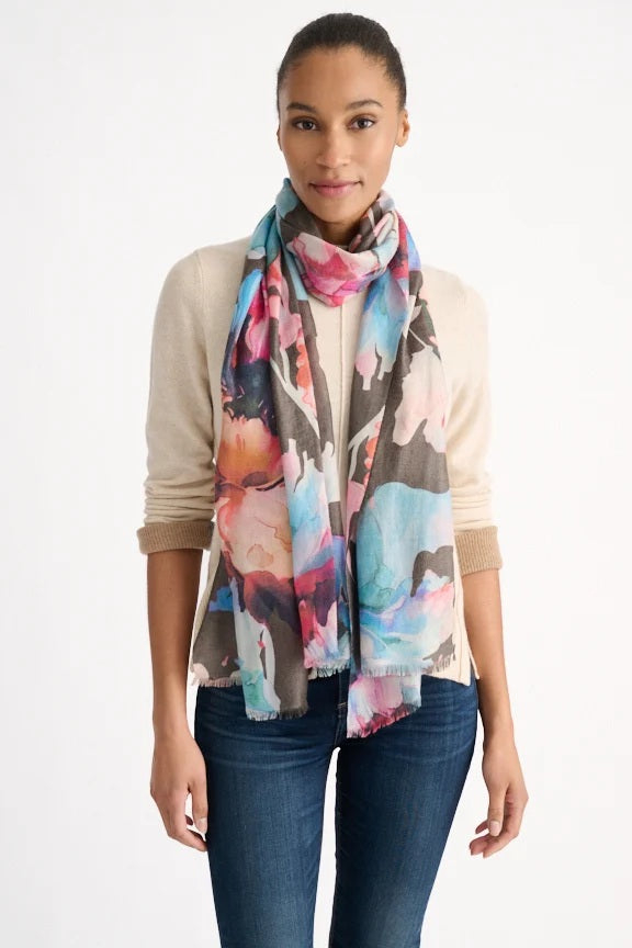 A person dressed in a cream sweater and blue jeans is shown with the KINROSS CASHMERE Highland Blooms Scarf in Chocolate from KINROSS wrapped around their neck, standing against a plain white background.