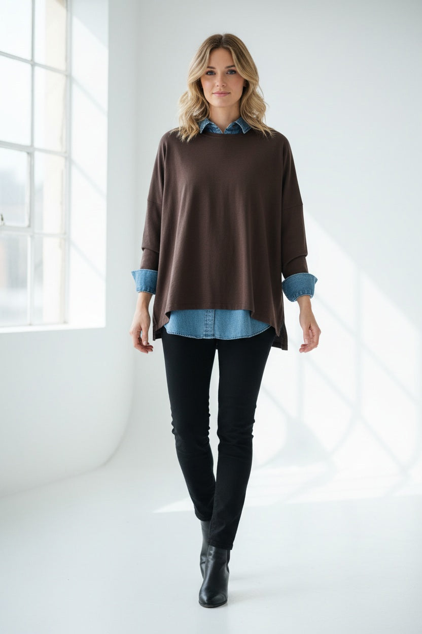 A woman with dark hair stands by a wooden door, wearing the MERSEA - Catalina Slub Tee in Espresso layered over a blue denim shirt and holding a black clutch. She looks at the camera, resting her arm on a red and white tiled wall.